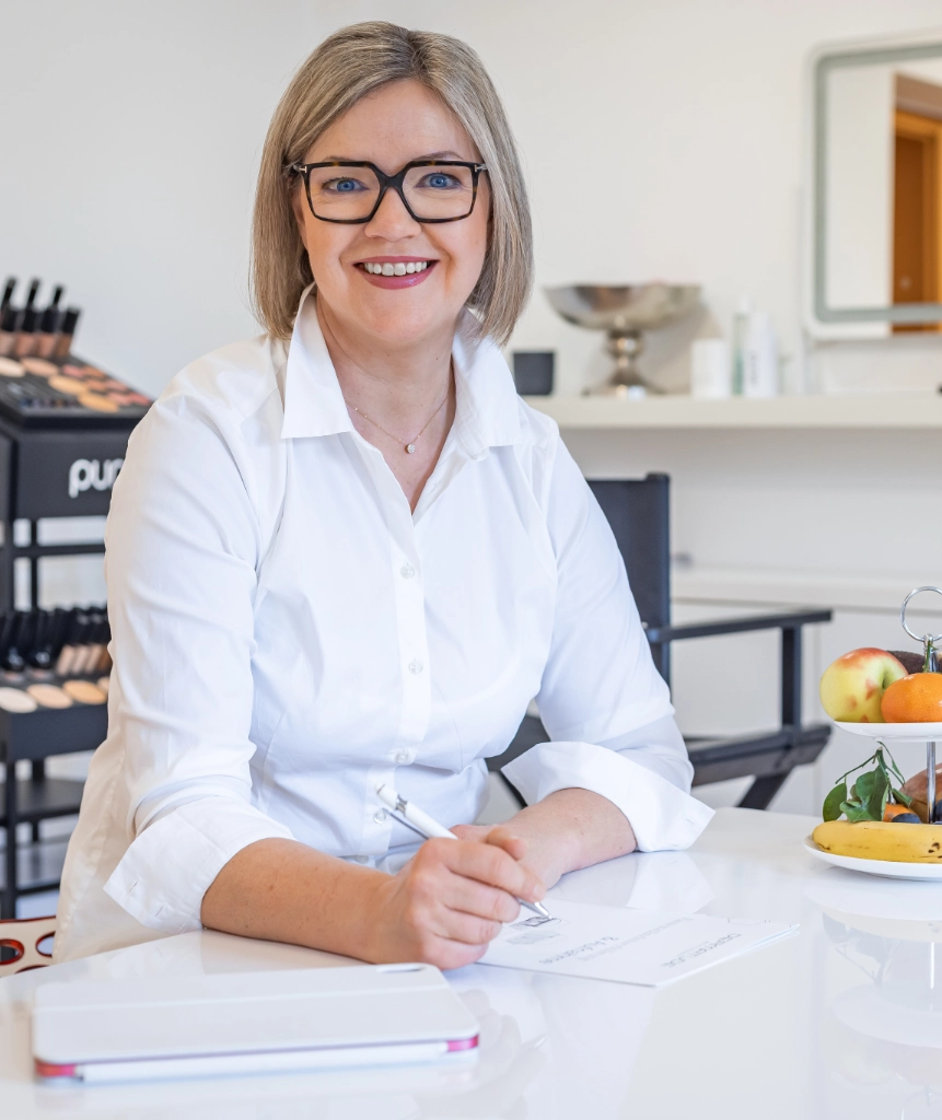 Lächelnde Frau mit Brille sitzt an einem Tisch und macht Notizen, im Hintergrund Kosmetikprodukte und ein Obstteller
