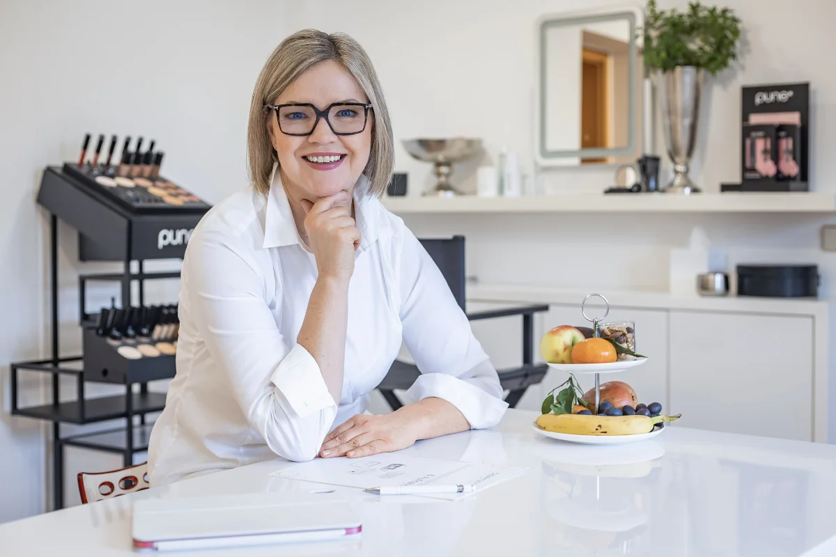 Lächelnde Frau mit Brille sitzt an einem Tisch in einem hellen Kosmetikstudio, neben ihr steht eine Etagere mit frischem Obst.