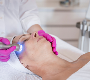 Nahaufnahme einer Frau, die auf einer Behandlungsliege liegt und eine Meta-Therapie-Behandlung erhält. Katrin Streit mit pinkfarbenen Handschuhen hält ein Gerät mit blauem Licht an die Schläfenpartie der Frau.