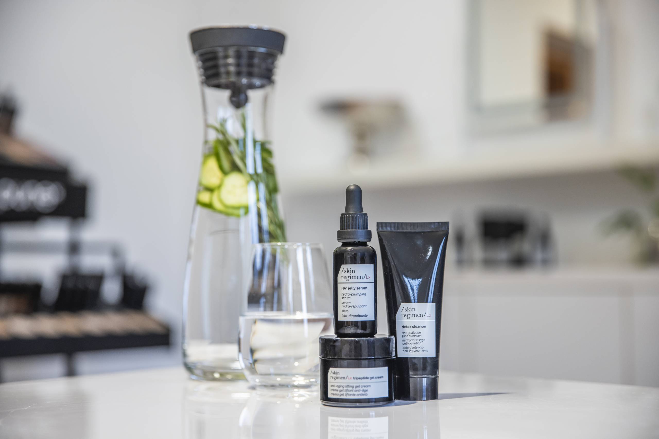 Hautpflegeprodukte der Marke Skin Regimen in einer schwarzen Flasche mit Pipette, einem schwarzen Tiegel und einer schwarzen Tube, arrangiert auf einem glänzenden Tisch. Daneben steht eine Glaskaraffe mit Gurkenscheiben und Kräutern im Wasser sowie ein gefülltes Wasserglas.